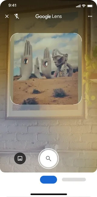 Sur un téléphone, l'application Google Lens est ouverte. Une notification indique à l'utilisateur qu'il peut appuyer sur le bouton de l'obturateur pour rechercher une image de grandes sculptures de mains en pierre dans un désert.