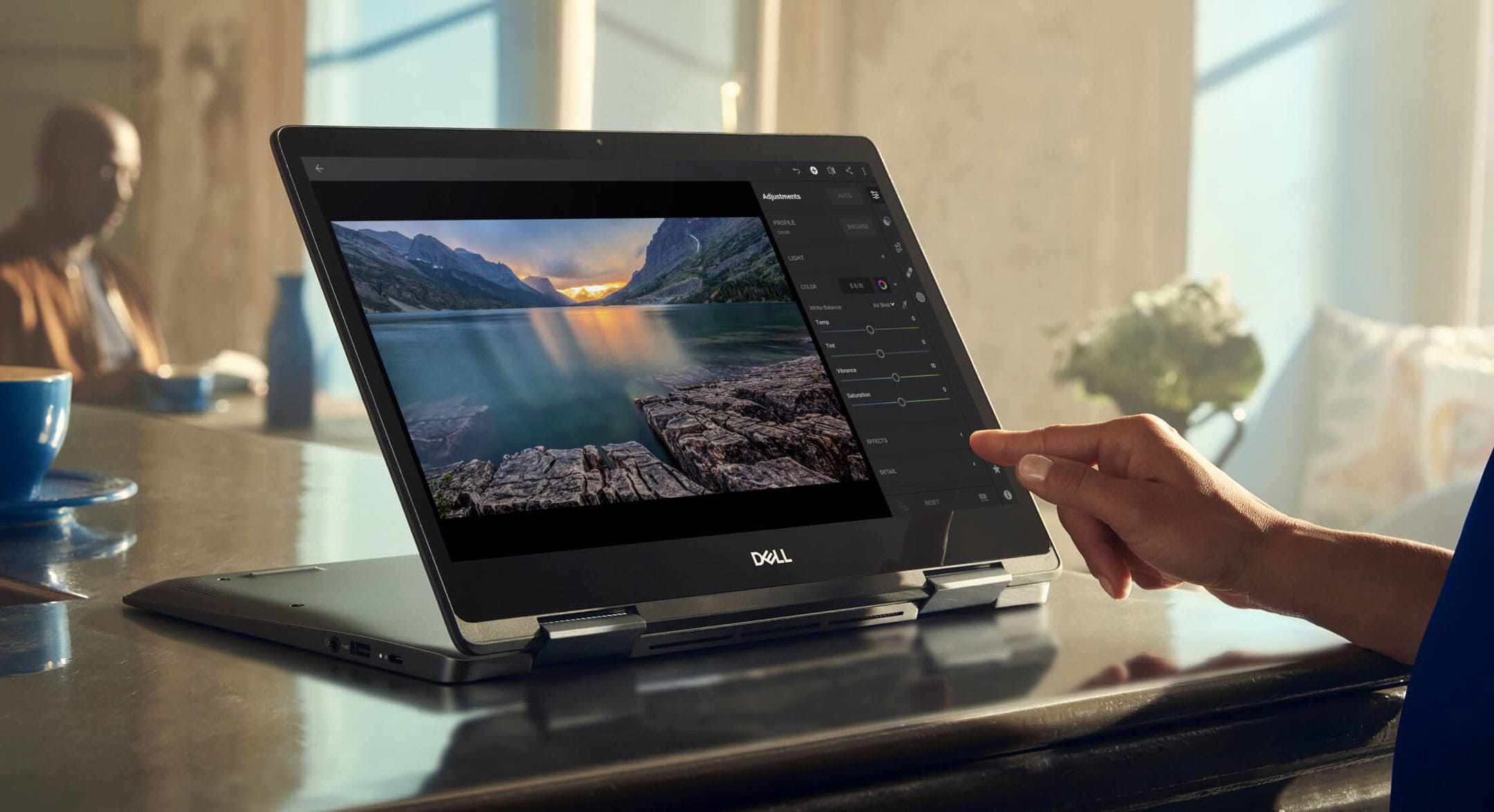 A woman in a coffee shop uses her Chromebook touchscreen to color correct some landscape pictures from a mountain trip.